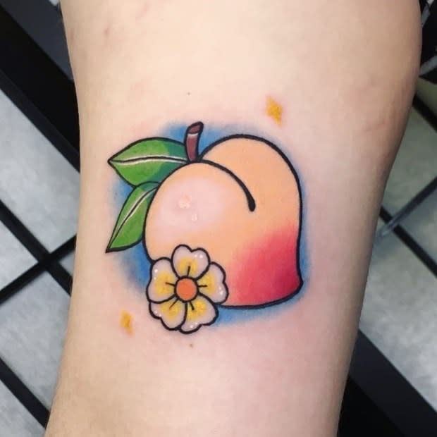 Tattoo of a peach with leaves and a yellow flower, highlighted with small yellow and blue sparkles